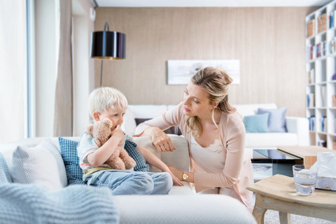 	Kleines Kind sitzt auf der Couch, Mutter kniet neben ihm und redet ihm gut zu