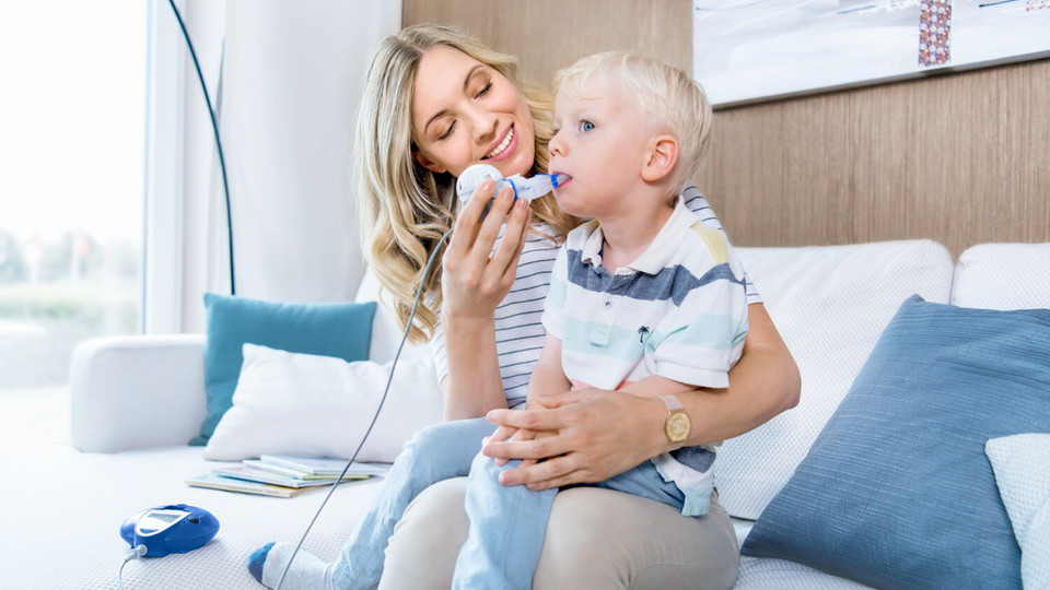 Mother sitting on the couch with her child on her lap and helping him inhale with the eFlow rapid inhalation device for cystic fibrosis