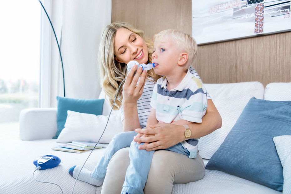 Mother sitting on the couch with her child on her lap and helping him inhale with the eFlow rapid inhalation device for cystic fibrosis