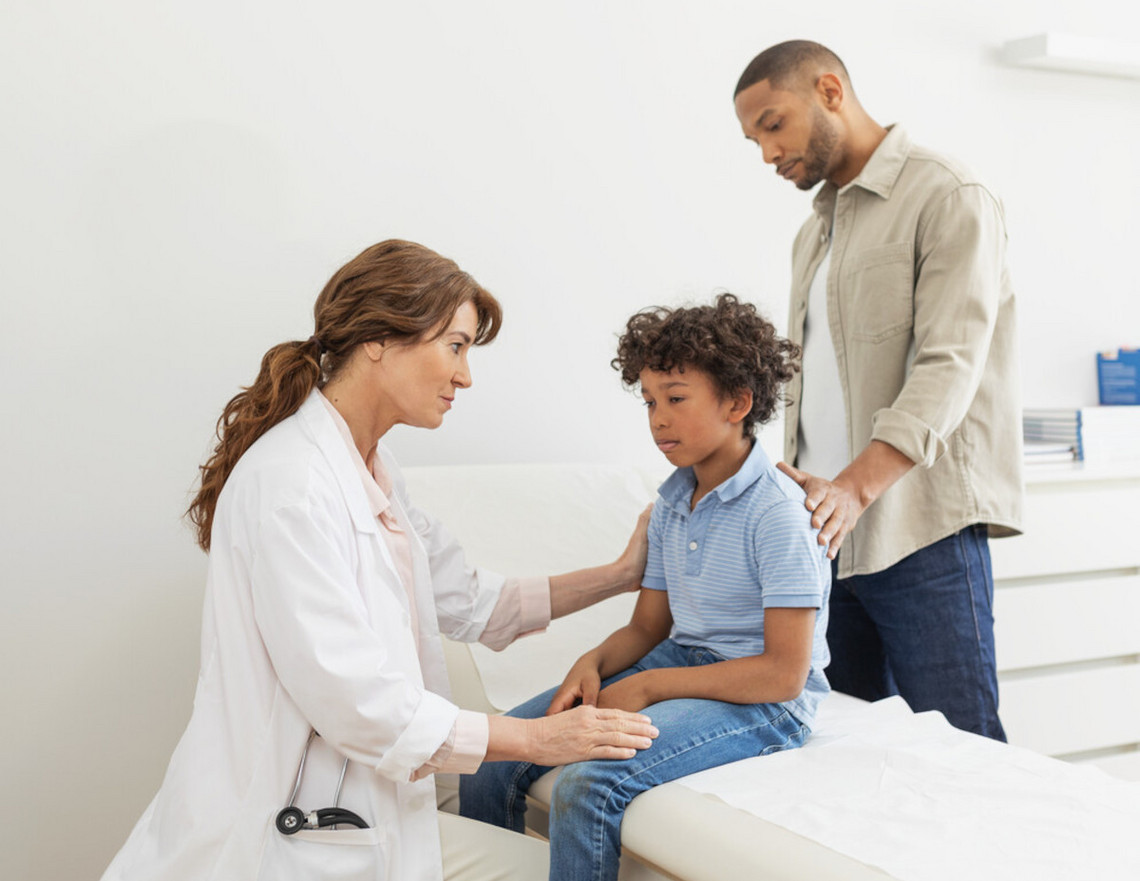 Child sits on the couch in front of a doctor, the father stands behind and has a hand on his back