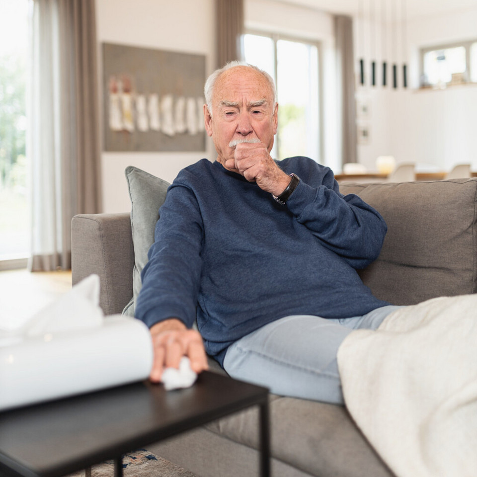 Älterer Mann sitzt hustend mit Faust vor dem Mund auf Sofa
