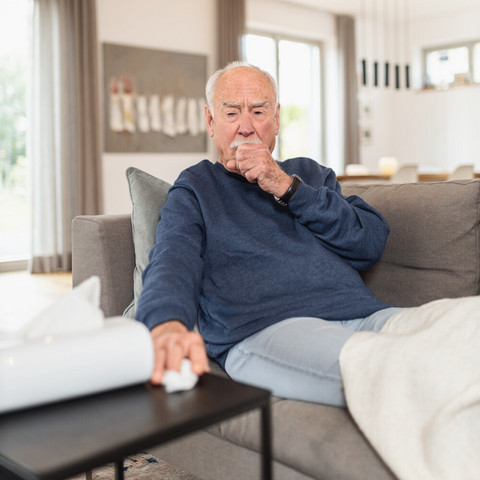 Älterer Mann sitzt hustend mit Faust vor dem Mund auf Sofa