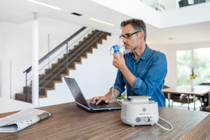 Mann inhaliert mit Inhalationssystem, während er am Tisch am Laptop arbeitet