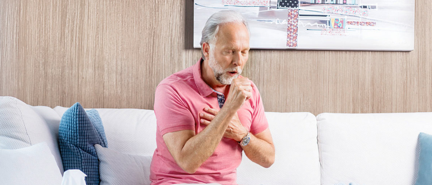 Ein älterer Mann mit grauem Bart und rosa Polohemd sitzt auf einem Sofa, hustet stark und hält sich die Hand vor die Brust. Neben ihm liegen Taschentücher und eine Brille.