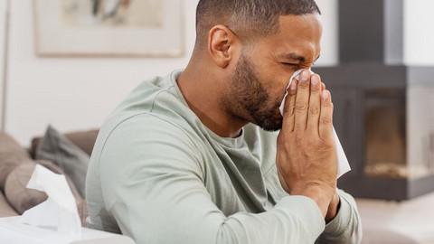 Junger Mann sitzt auf Couch und schnäuzt sich die Nase