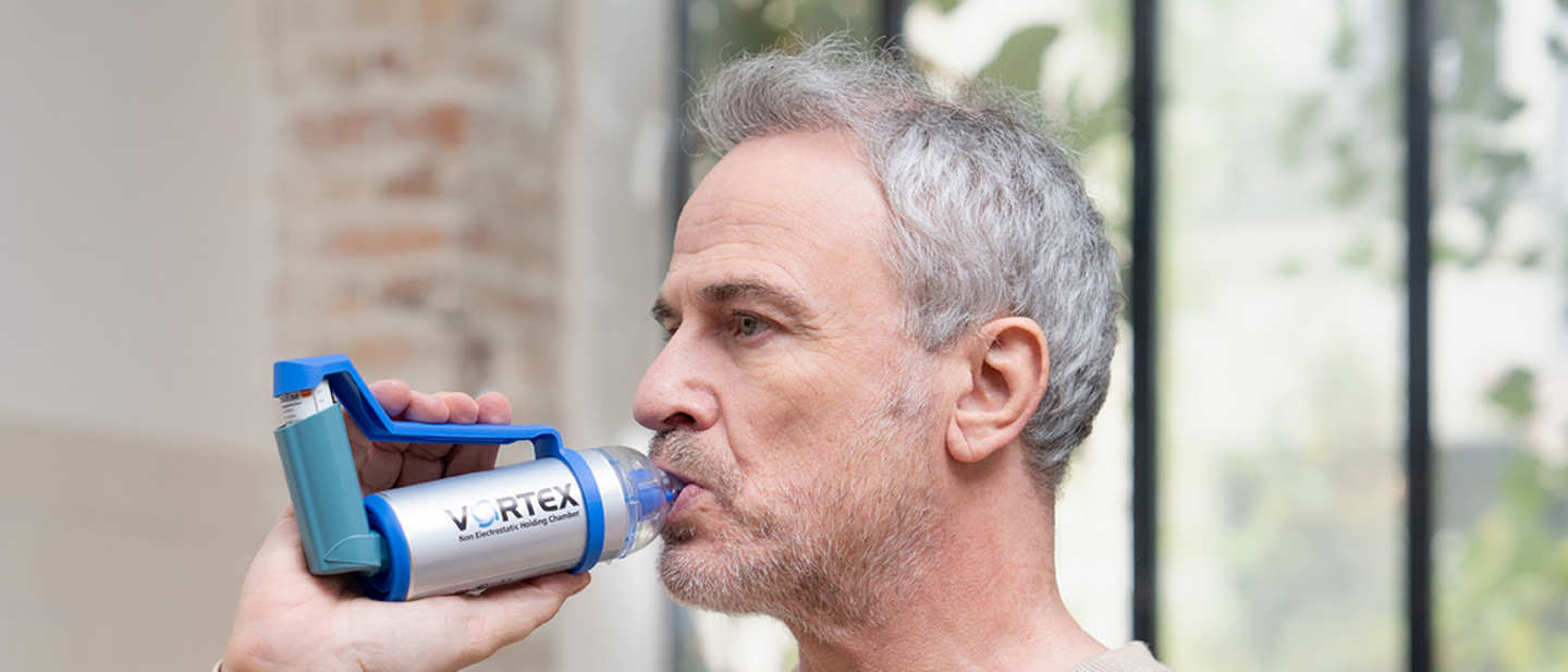 Inhalieren mit Asthmaspray Erwachsene Person inhaliert ein Asthmaspray mit VORTEX Vorschaltkammer vor einem Fenster in einem hellen Raum.