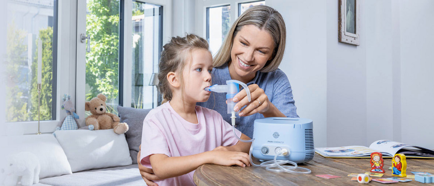 A mother helps her young daughter inhale with the PARI COMPACT2 inhaler. The girl holds the mouthpiece in her mouth. Both are sitting at a round wooden table in a bright, cheerfully furnished room with large windows.