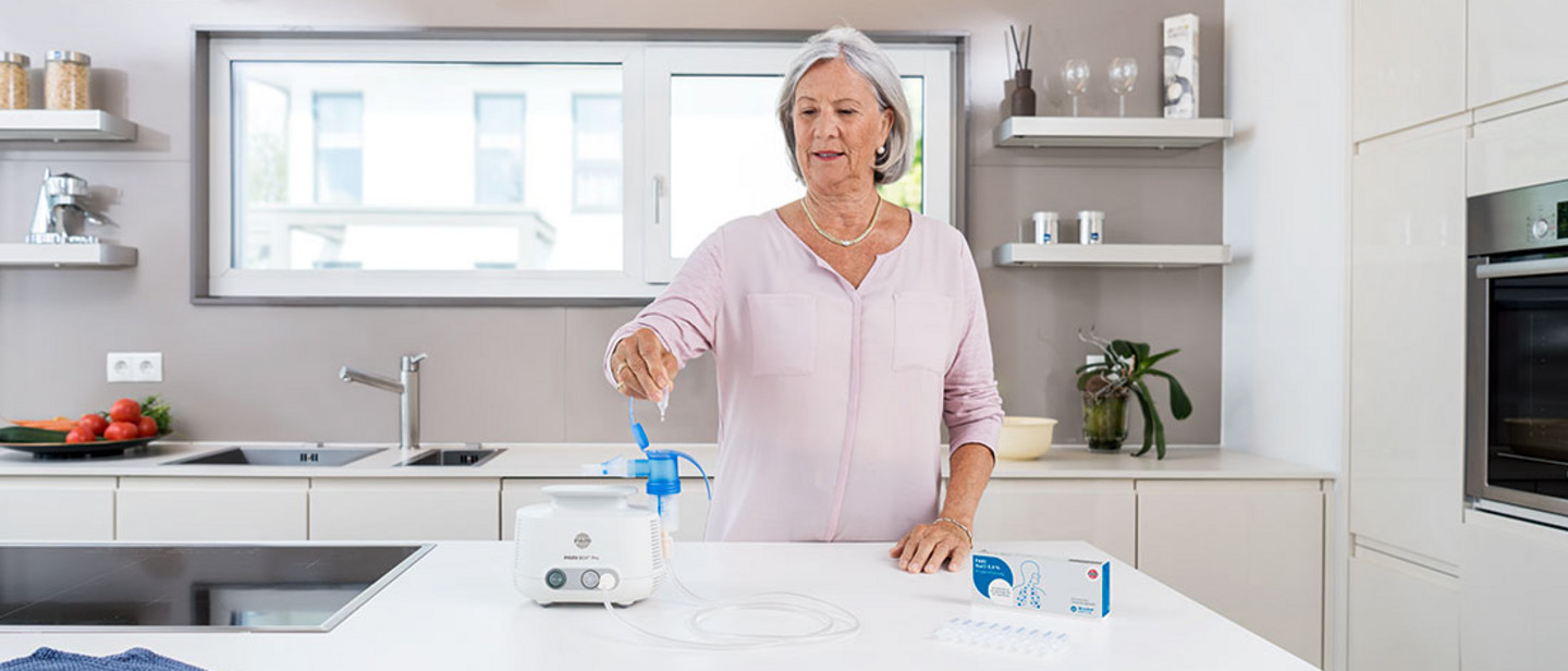 Une femme utilise un inhalateur PARI dans une cuisine moderne pour suivre un traitement respiratoire ; des ampoules de médicaments et des accessoires sont à portée de main.