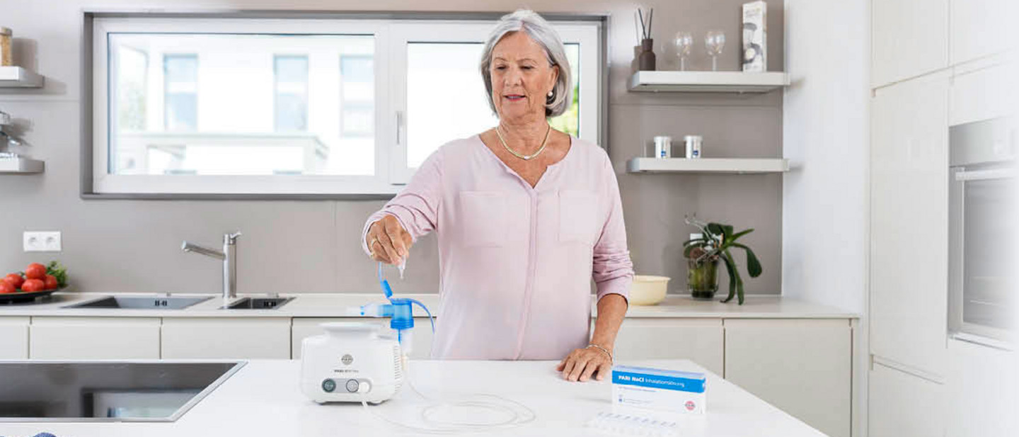 Ein Frau nutzt einen PARI-Inhalator in einer moderner Küche zur Atemtherapie, Medikamentenampullen und Zubehör liegen bereit.