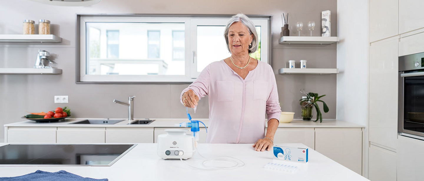 Ein Frau nutzt einen PARI-Inhalator in einer moderner Küche zur Atemtherapie, Medikamentenampullen und Zubehör liegen bereit.