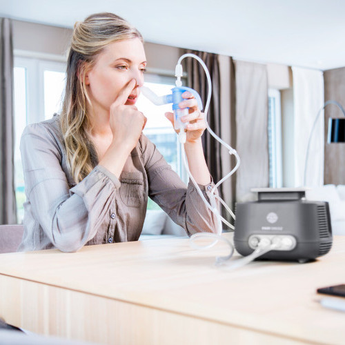 Frau sitzt im Wohnzimmer am Tisch und hält Vernebler an ihr Nasenloch, Inhalationsgerät steht auf dem Tisch