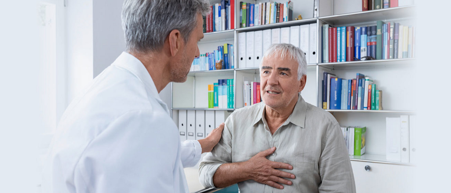 An elderly man sits in a doctor's office, holds his hand on his chest and talks to a doctor in a white coat who places a reassuring hand on his shoulder.