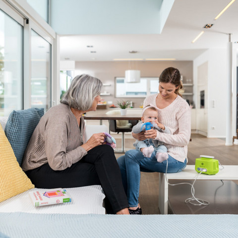 Junge Mutter sitzt mit Baby auf dem Schoß auf dem Couch-Tisch und hält ihm Vernebler mit Maske vors Gesicht, PARI BOY Junior steht auf dem Tisch, Oma sitzt auf der Couch und schaut zu