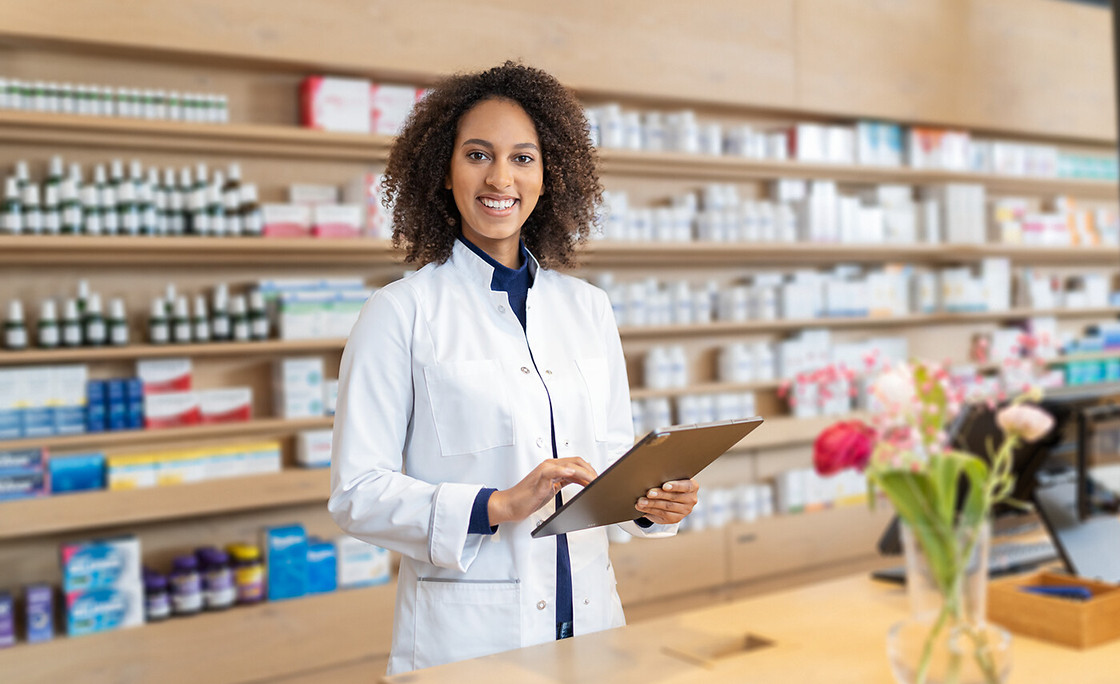 Apothekerin im weißen Kittel steht in einer Apotheke und nutzt ein Tablet; im Hintergrund Regale mit Medikamenten.