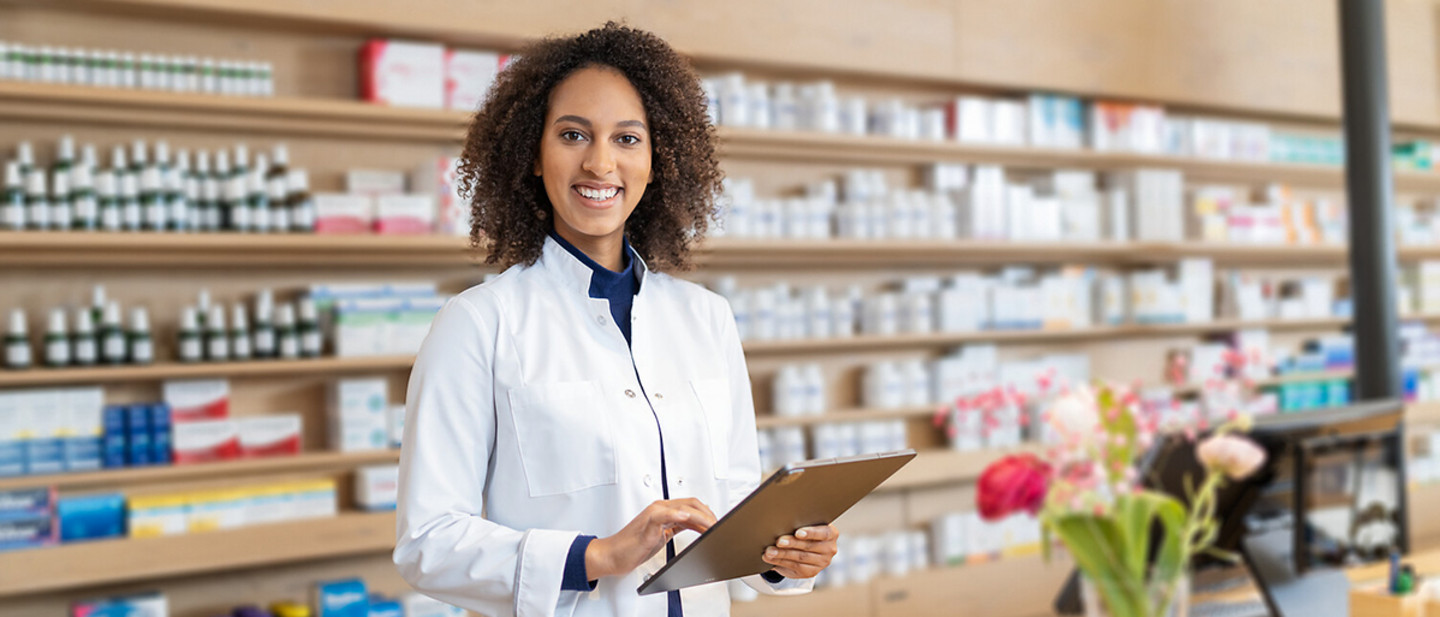 Apothekerin im weißen Kittel steht in einer Apotheke und nutzt ein Tablet Apothekerin im weißen Kittel steht in einer Apotheke und nutzt ein Tablet; im Hintergrund Regale mit Medikamenten.