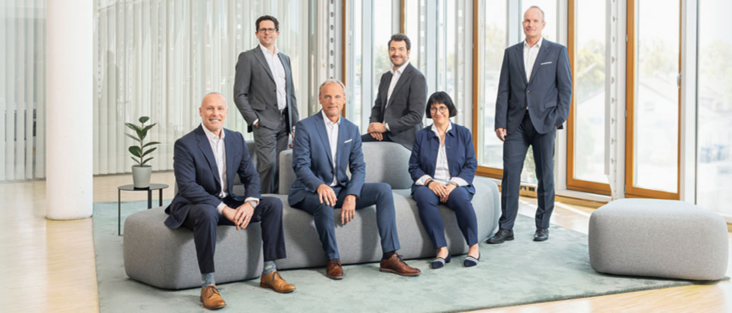 Cinq hommes et une femme en tenue professionnelle sont debout et assis dans un espace moderne et lumineux, sur des sièges gris.