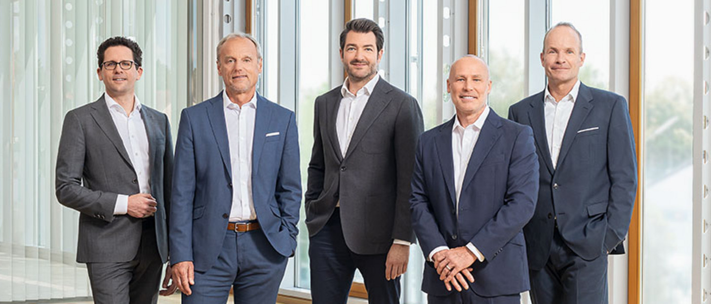 Fünf Männer und eine Frau in Businesskleidung stehen und sitzen in einem modernen, lichtdurchfluteten Raum auf grauen Sitzmöbeln.