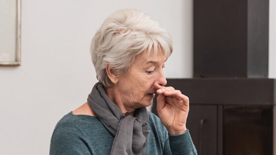 Ältere Frau sitzt im Wohnzimmer und räuspert sich