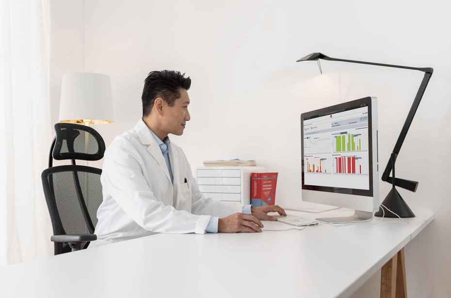 Doctor in a white coat sitting at a desk and analyzing data charts on a large desktop computer in a modern, well-lit office.