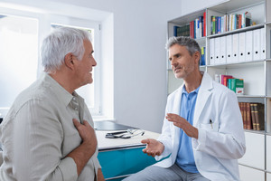 Älterer Mann sitzt mit Hand auf Brust vor Arzt, der ihm etwas erklärt