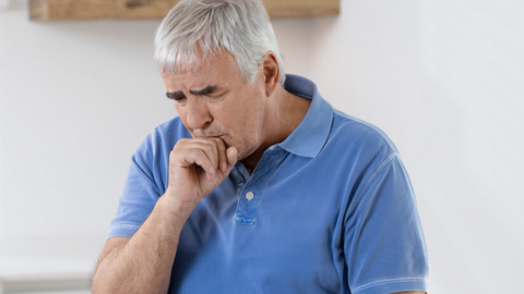 Älterer Mann sitzt in der Küche am Tisch, er hustet und hält sich die Faust vor den Mund