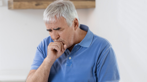 Älterer Mann sitzt in der Küche am Tisch, er hustet und hält sich die Faust vor den Mund