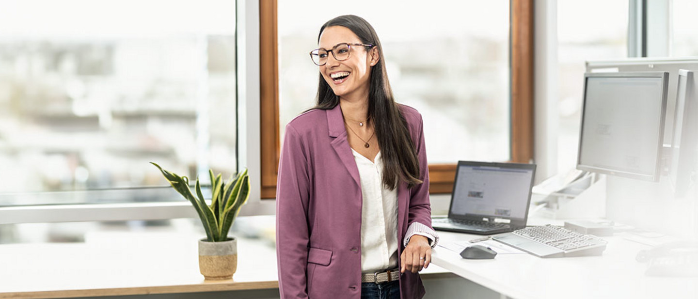 Lächelnde Frau mit Brille und lila Blazer steht in einem modernen Büro neben einem Schreibtisch mit mehreren Monitoren und einem Laptop.