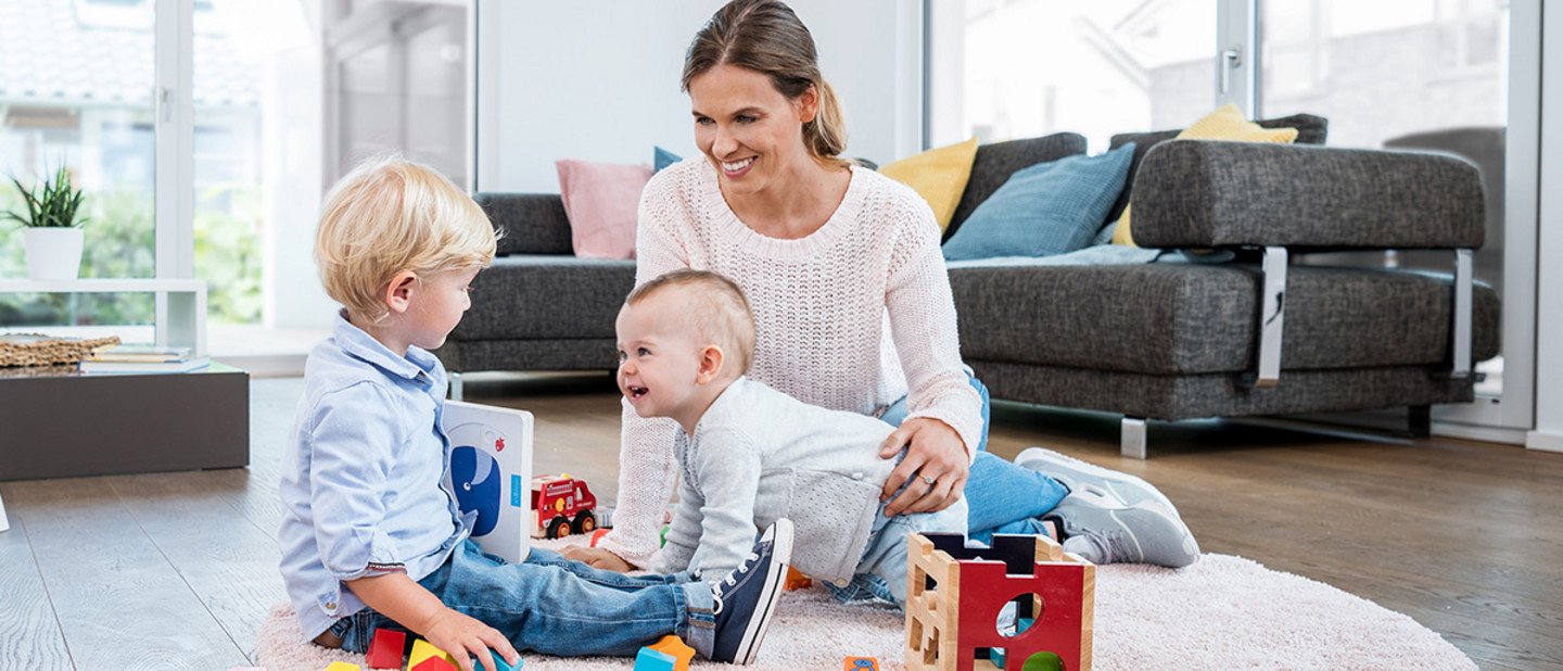 Erwachsene Person spielt mit zwei Kleinkindern auf rosa Teppich mit Lernspielzeug in hellem, gemütlichem Wohnzimmer.