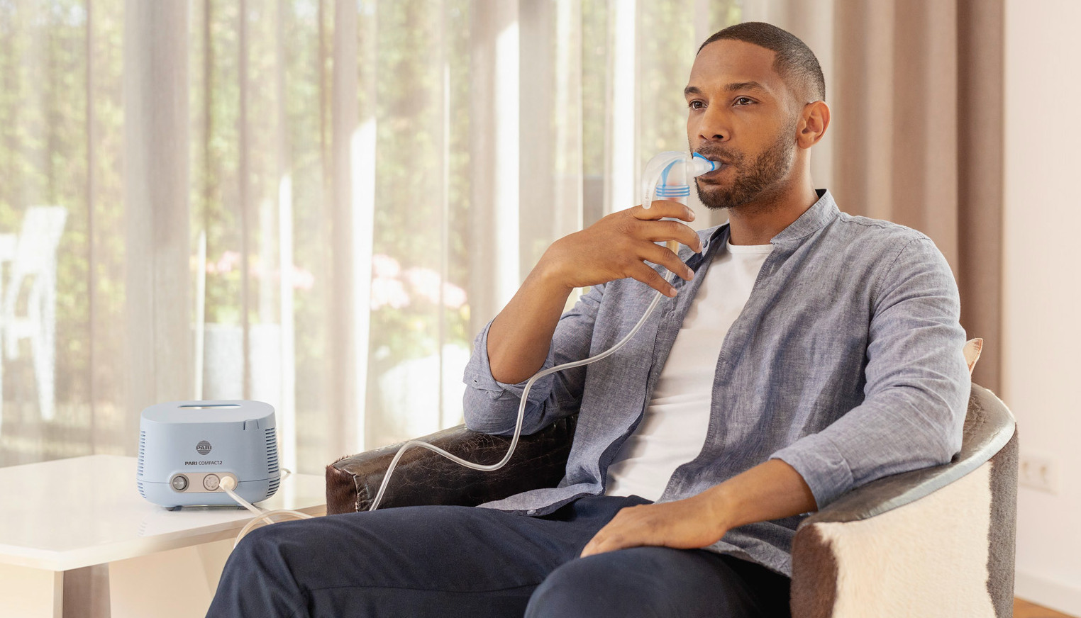 A man sits relaxed in an armchair and inhales using the PARI COMPACT2 inhalation device.