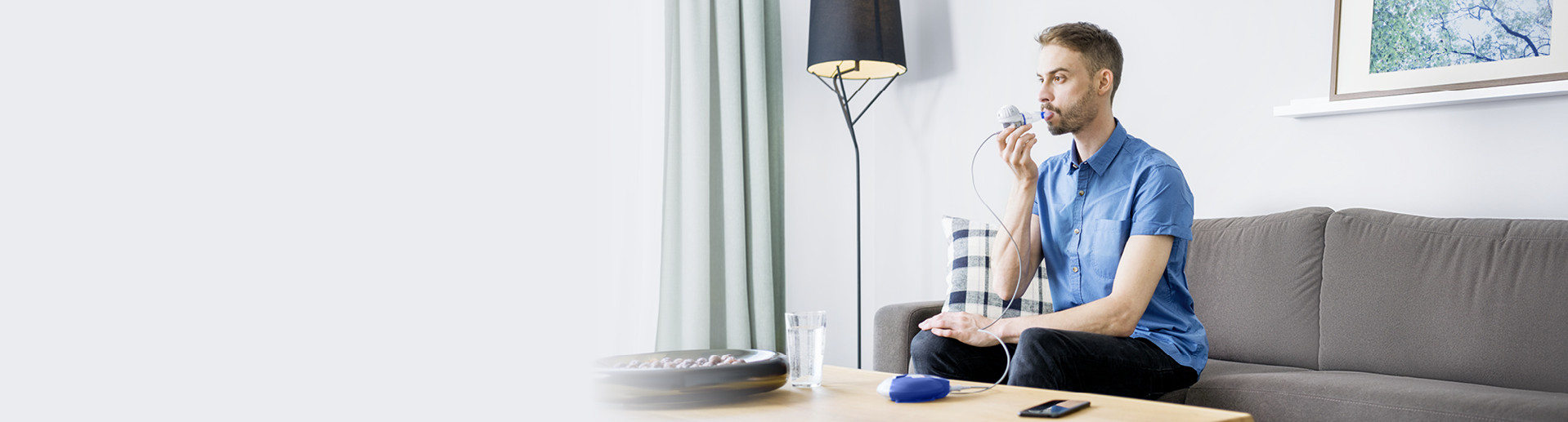 Ein Mann mit blauem Hemd sitzt entspannt auf einem grauen Sofa in einem modern eingerichteten Wohnzimmer. Er hält einen eFlow rapid Vernebler mit Mundstück und nutzt es für seine Therapie. Auf dem Holztisch vor ihm liegen ein Smartphone und der Controller.