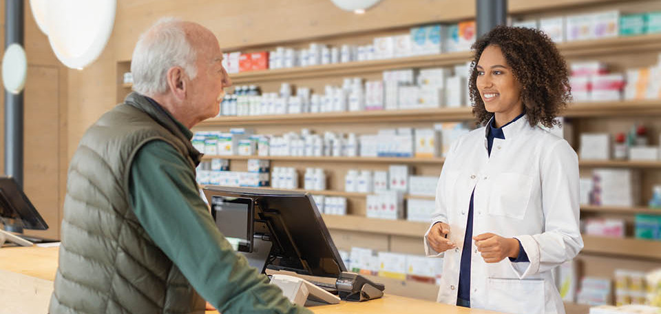 In einer Apotheke steht eine Apothekerin hinter der Theke und erklärt die Funktionsweise des PARI COMPACT2 Inhaliergerätes, das vor ihr auf der Theke steht. Lächelnd legt sie dabei die rechte Hand auf das Gerät. Rechts neben Ihr steht eine Vase mit Blumen. Die Apotheke ist modern, hell und freundlich.