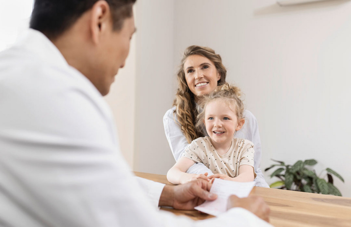 Lächelnde Mutter sitzt mit ihrer Tochter auf dem Schoß an einem Tisch, während ein Arzt ihnen gegenüber ein Blatt in den Händen hält.
