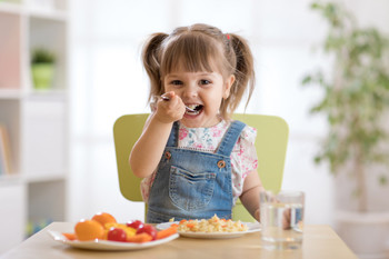 Kleines Kind sitzt an Tisch und isst mit Freude, zwei Teller und ein Glas Wasser stehen vor ihm