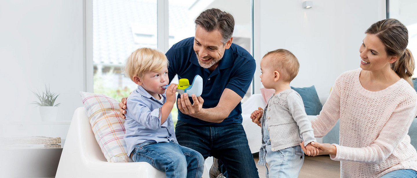 A father helps his little boy use an inhaler while the mother holds the hand of a second younger child and watches, smiling.
