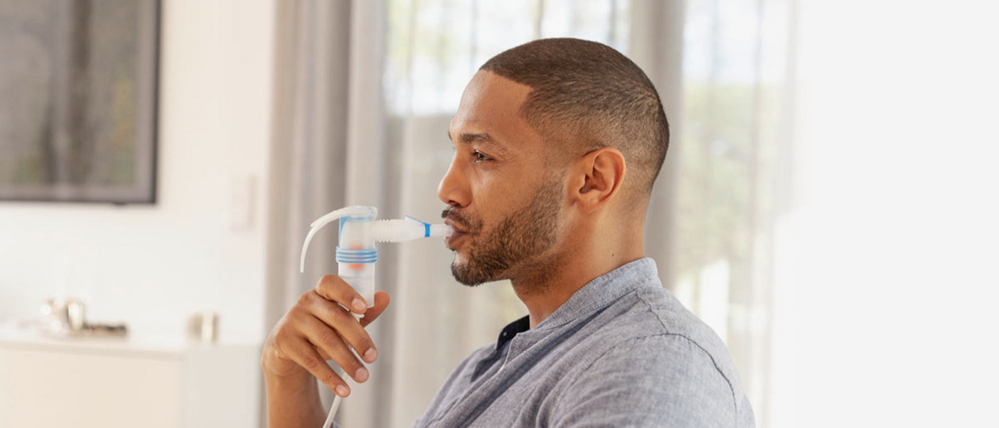 Ein Mann mit kurzem Bart und kurzem Haar sitzt entspannt in einem hellen Wohnzimmer und inhaliert mit einem Vernebler. Er hält das Mundstück des Inhalationsgeräts an seinen Mund und atmet ein.