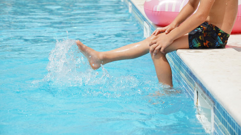 Junge sitzt am Beckenrand, die Beine im Wasser, Kopf ist nicht im Bild