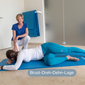 Junge Frau liegt auf einer Matte am Boden und macht die Übung Brust-Dreh-Dehn-Lage, während die Physiotherapeutin sie anleitet