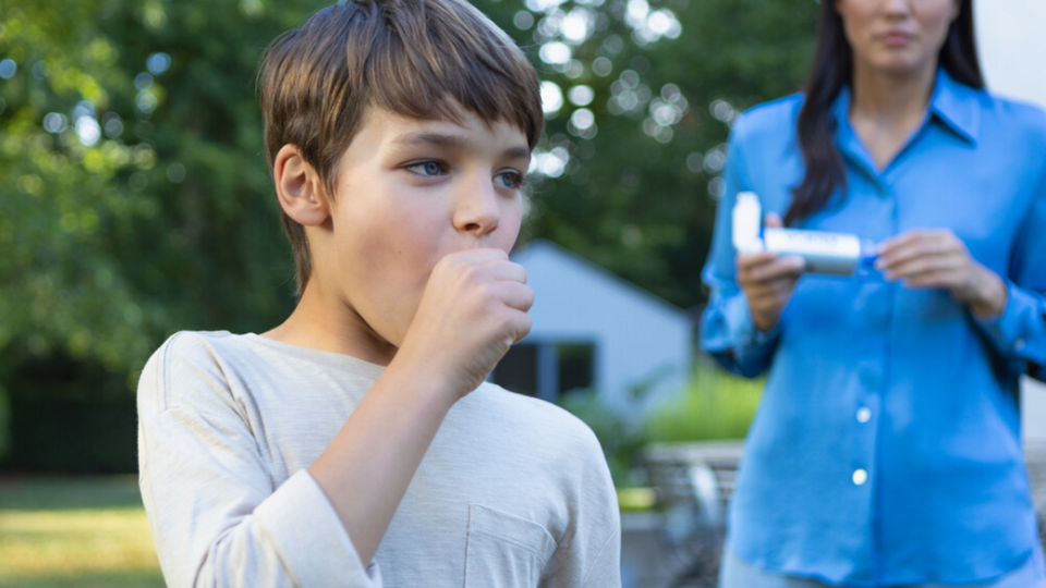 Junge hustet und hält sich die Faust vor dem Mund, im Hintergrund ist Mutter mit Asthmaspray zu sehen