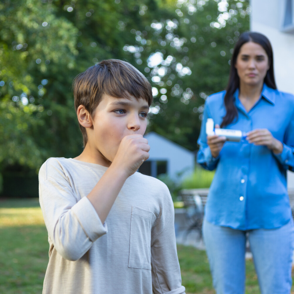 Junge hustet und hält sich die Faust vor dem Mund, im Hintergrund ist Mutter mit Asthmaspray zu sehen