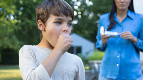Junge hustet und hält sich die Faust vor dem Mund, im Hintergrund ist Mutter mit Asthmaspray zu sehen