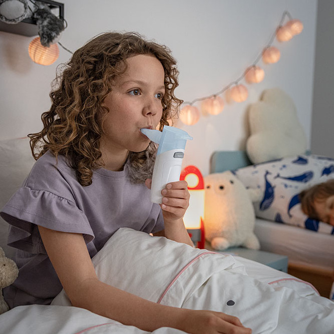 Kind sitzt im Bett und inhaliert mit einem PARI BOY Go Kind sitzt im Bett und inhaliert mit einem PARI BOY Go, während ein weiteres Kind im Hintergrund schläft.
