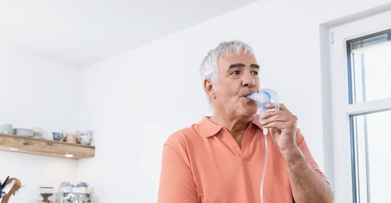 Elderly man uses a nebulizer for respiratory therapy in a bright kitchen with natural light and household items in the background.