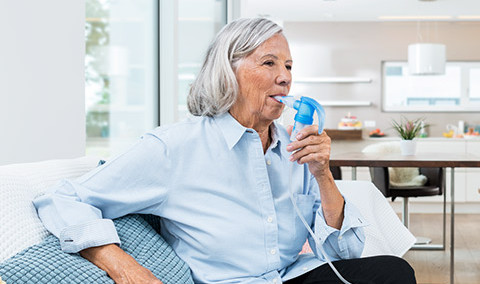 Elderly woman sitting on a couch using a nebulizer device for respiratory treatment in a bright, modern living room.
