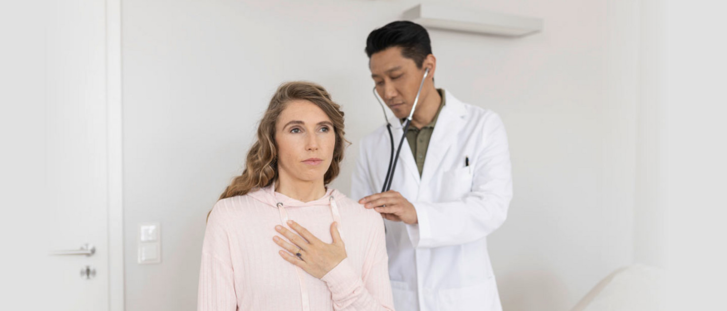Ein Arzt in weißem Kittel hört mit einem Stethoskop den Rücken einer Frau ab. Die Frau sitzt aufrecht und hält eine Hand auf ihre Brust.