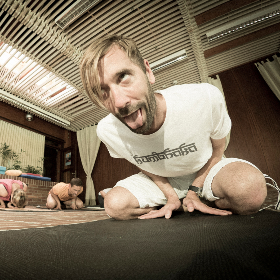 Mann macht Grimasse und kniet auf Yogamatte, die Hände zwischen seinen Beinen abgestützt