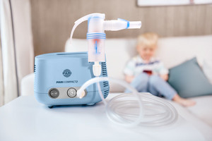 An inhaler can be seen in the foreground, a child is sitting on the sofa in the background