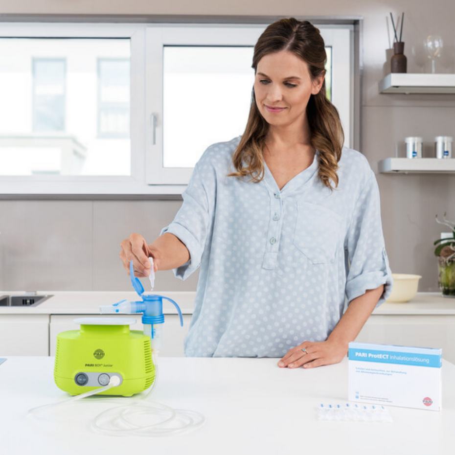 Young woman standing at the kitchen table filling the PARI BOY Junior inhalation device with inhalation solution