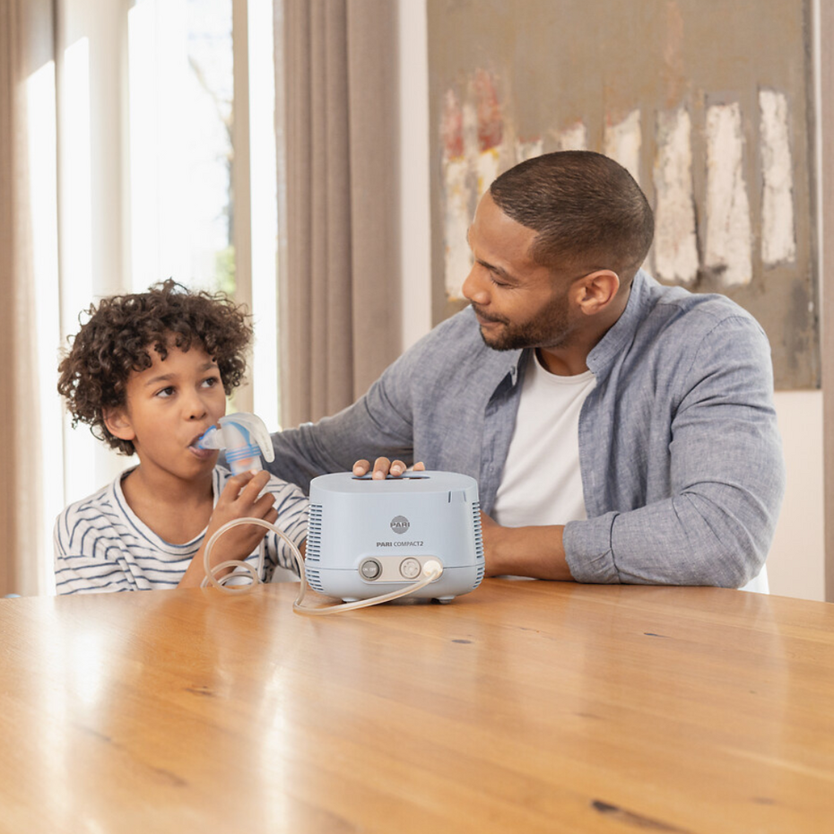 Junge inhaliert am Tisch mit dem PARI COMPACT2 Inhalationsgerät, Vater sitzt lächelnd daneben
