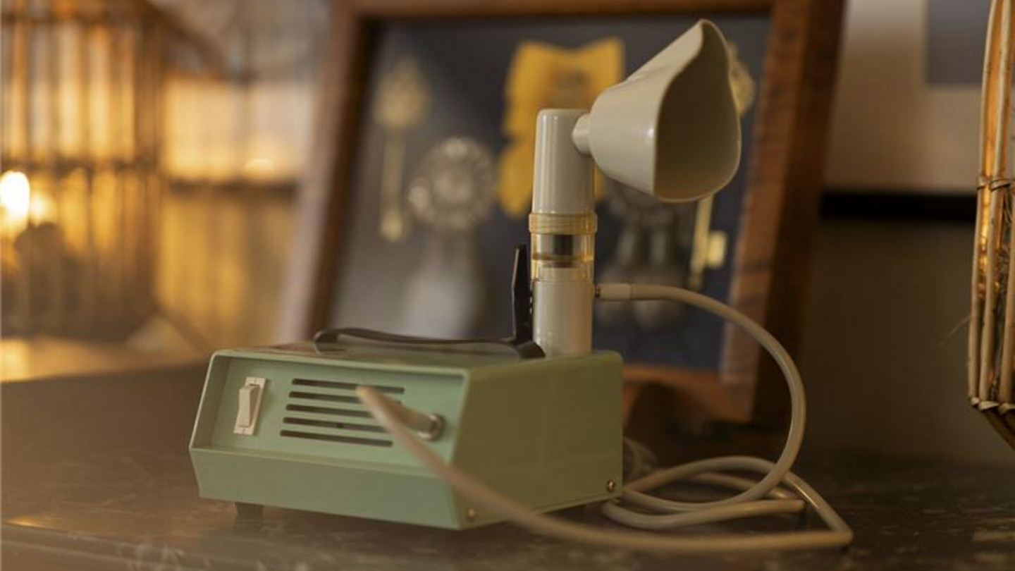 A vintage inhalation device with mouthpiece and hose stands on a table in front of a framed picture.