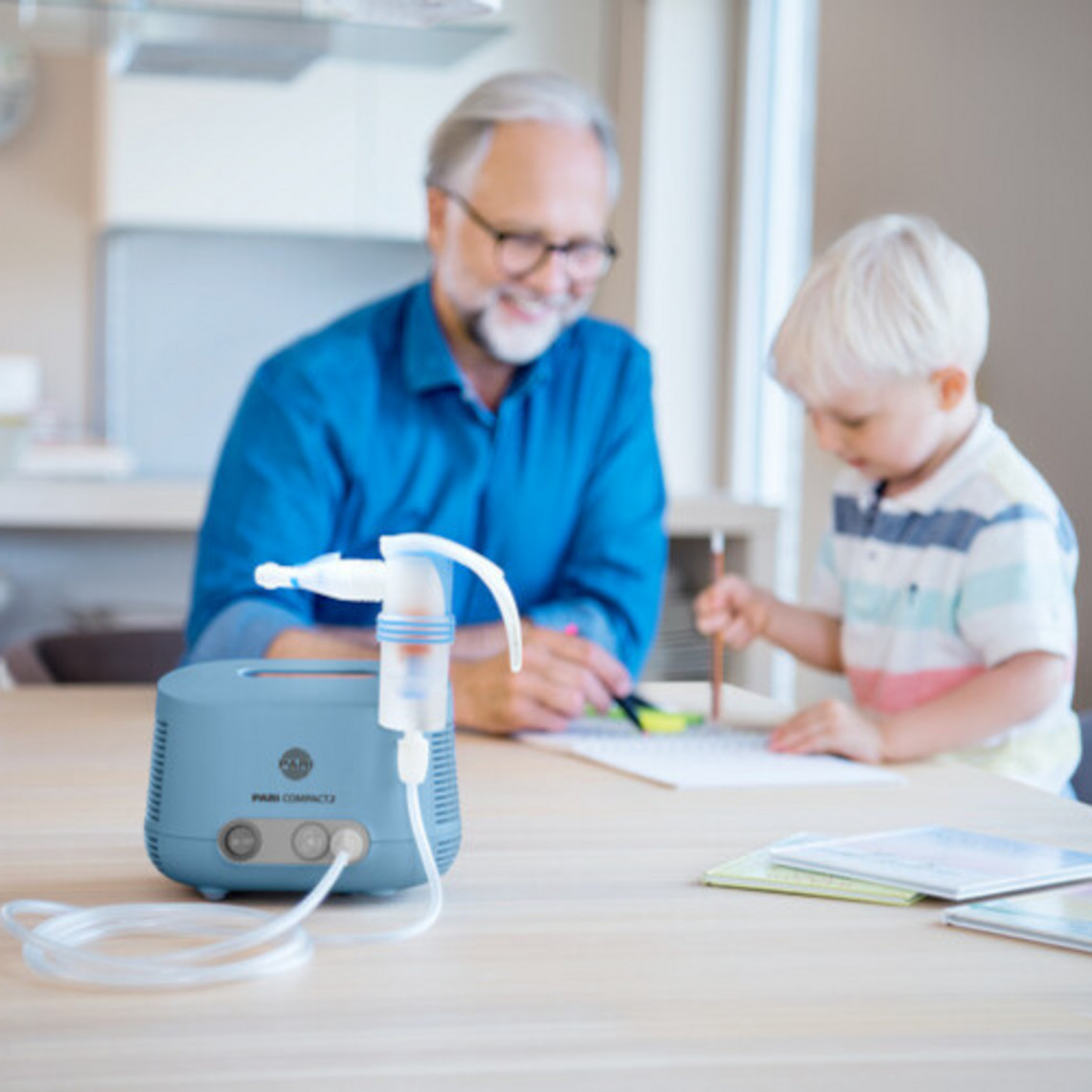 Opa spielt im Hintergrund mit Enkel am Tisch, im Vordergrund steht ein Inhalationsgerät mit Vernebler